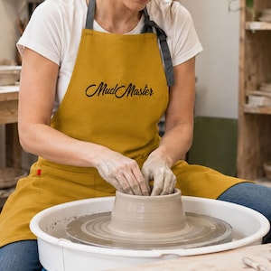 May include: A mustard yellow apron featuring the text Mud Master in black script lettering. The apron has dark grey neck straps and a front pocket with a small red tab. It is worn over a white t-shirt while shaping clay on a pottery wheel.