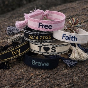 May include: A collection of woven friendship bracelets with embroidered text and tassels. The bracelets are in various colors, including pink, black, and white. The text includes words like "Free," "Faith," and "Brave," along with a date and initials.