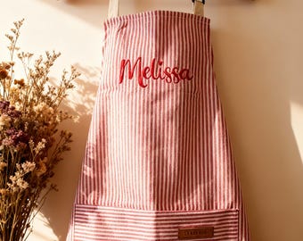 May include: A red and white vertically striped apron hanging on a wooden hanger. The apron features the name Melissa embroidered in red script across the chest and a large front pocket with a small rectangular brown leather-like brand label.
