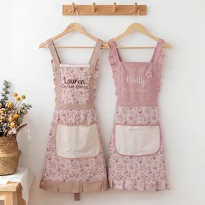 Op de afbeelding: Twee roze bloemen schorten hangen aan houten hangers. Eén schort heeft de tekst "Lauren Cookie Queen" en de andere heeft de tekst "Molly Mum's Helper". Beide schorten hebben een grote voorzak en gefranjerde randen.
