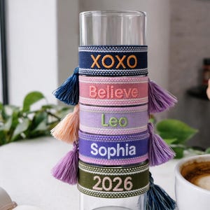 Könnte beinhalten: Ein Stapel bunter, bestickter Stoffarmbänder mit Quasten. Die Armbänder sind marineblau, rosa, lavendelfarben, lila und olivgrün. Jedes Armband hat einen gestickten Text: "XOXO", "Believe", "Leo", "Sophia" und "2026".