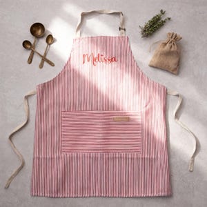 May include: A red and white pinstriped apron featuring the name Melissa embroidered in red script on the chest. The apron includes a large front pocket with a small rectangular label, a neck strap, and two side ties for securing the garment.