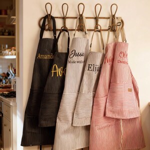 Puede incluir: Cinco delantales a rayas con bordados personalizados colgados en un perchero de pared de madera. Los delantales presentan varios colores, incluyendo negro, gris y rojo con rayas blancas, cada uno equipado con bolsillos delanteros y tiras ajustables.