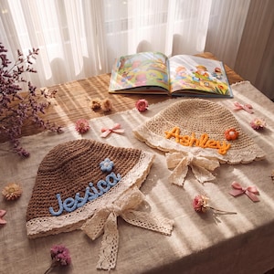 May include: Two straw sun hats with personalized names, "Jessica" and "Ashley", are displayed on a table. The brown hat has a blue name, while the beige hat has an orange name. Both hats have a lace trim and bow.