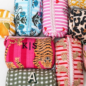 May include: A collection of rectangular quilted fabric pouches featuring various patterns including stripes, floral prints, leopard spots, and tiger motifs. Several pouches are personalised with embroidered letters or names in contrasting thread colours.