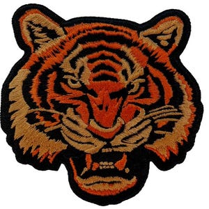 May include: Embroidered patch featuring a tiger's head with an open mouth, showcasing sharp teeth. The design uses orange, black, and tan thread, with a black border. The tiger's face is detailed with stripes and facial features.
