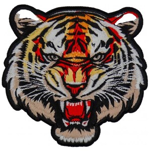 May include: Embroidered patch featuring a detailed tiger head with an open mouth, showcasing sharp teeth. The design incorporates shades of white, black, tan, yellow, and red, creating a fierce and striking image. The patch is likely for application on clothing or accessories.