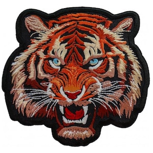 Puede incluir: Parche bordado con la cara de un tigre con rayas naranjas y negras, ojos azules y boca abierta. El parche tiene un borde negro, diseñado para ser aplicado en ropa o accesorios.