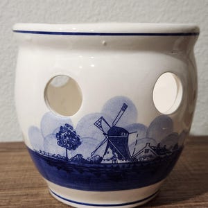 May include: A white ceramic pot with a blue Dutch windmill design. The pot has three circular holes and a blue band around the base. The rim is also trimmed with a thin blue line. The design features a windmill, trees, and houses.