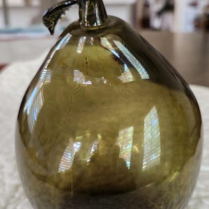 May include: An olive-green glass pear-shaped ornament. The pear has a stem and a curved, leaf-like detail. The glass reflects light, creating highlights and shadows. The pear is sitting on a white, textured surface.