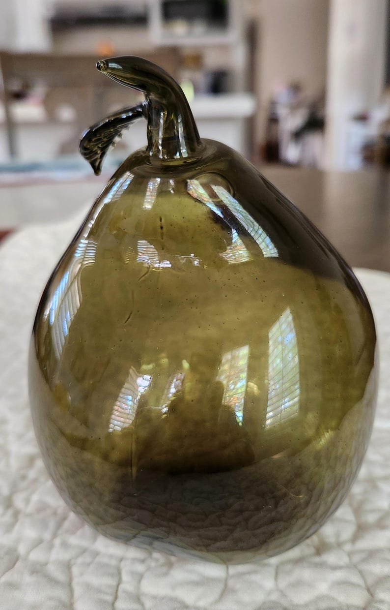 May include: An olive-green glass pear-shaped ornament. The pear has a stem and a curved, leaf-like detail. The glass reflects light, creating highlights and shadows. The pear is sitting on a white, textured surface.