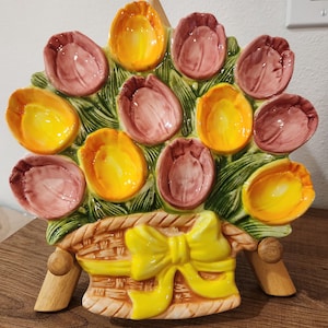 May include: A ceramic tulip-shaped serving dish. The dish features a basket base with a yellow bow, supporting a bouquet of colorful tulips in shades of yellow and pink. The leaves are green, and the dish is supported by wooden legs.