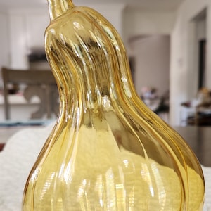 May include: A decorative, amber-colored glass gourd with a curved stem. The gourd has a ribbed texture and a glossy finish, reflecting light. The design is reminiscent of autumn decor.