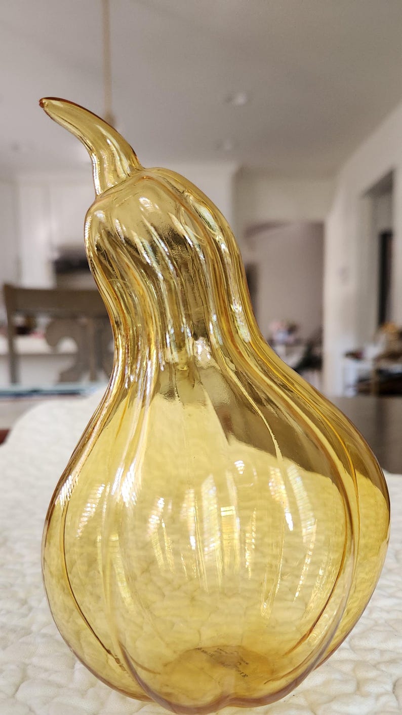 May include: A decorative, amber-colored glass gourd with a curved stem. The gourd has a ribbed texture and a glossy finish, reflecting light. The design is reminiscent of autumn decor.