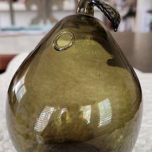 May include: An olive-green glass pear-shaped ornament with a stem and leaf detail. The glass has a textured surface and a small circular opening. The pear reflects light, creating highlights and shadows.