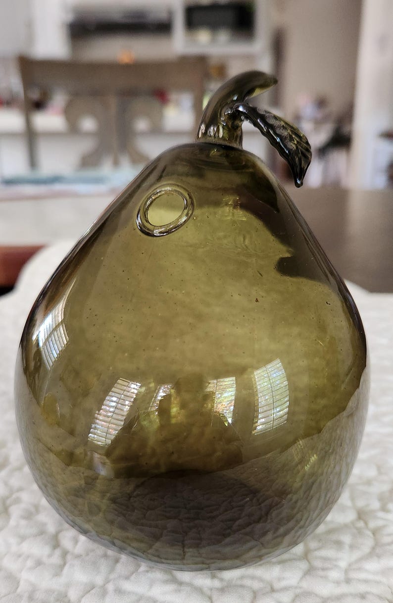 May include: An olive-green glass pear-shaped ornament with a stem and leaf detail. The glass has a textured surface and a small circular opening. The pear reflects light, creating highlights and shadows.