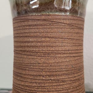 May include: A handcrafted ceramic vase with a textured, wrapped design. The upper portion is glazed in a mottled green, transitioning to a band of brown, wrapped texture. The base is a darker brown. This unique piece is suitable for home decor.