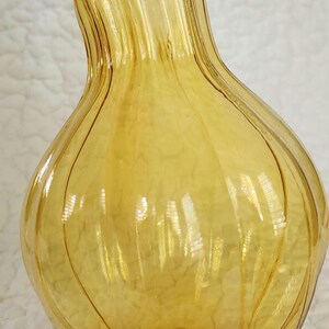 May include: A decorative, amber-colored glass vase with a bulbous body and a slender, curved neck. The vase has vertical ridges and a small opening at the top. The vase is approximately 12 inches tall.