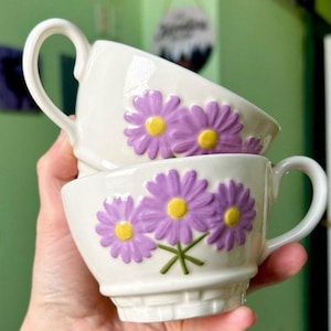 May include: Two white ceramic teacups with purple daisy designs and yellow centers. The cups have curved handles and a basket weave pattern at the base. The daisies are raised and add a 3D effect to the cups.
