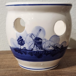 May include: A white ceramic pot with a blue Dutch windmill design. The pot has two circular openings and a blue band around the base. The design features a windmill, trees, and houses, all in shades of blue.