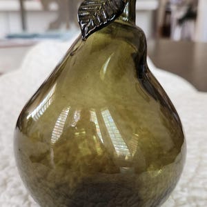 May include: An olive green glass pear ornament. The pear-shaped body has a textured surface, and a stem with a leaf detail extends from the top. The glass has a slight transparency. A decorative item.