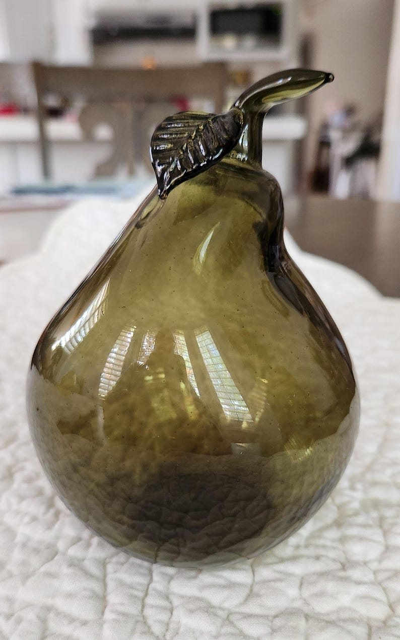May include: An olive green glass pear ornament. The pear-shaped body has a textured surface, and a stem with a leaf detail extends from the top. The glass has a slight transparency. A decorative item.