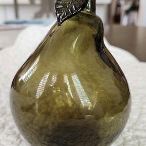 May include: Olive green glass pear-shaped decorative object. The pear has a textured surface and a stem with a leaf detail. The glass is translucent, allowing light to pass through. This decorative piece is suitable for home decor.
