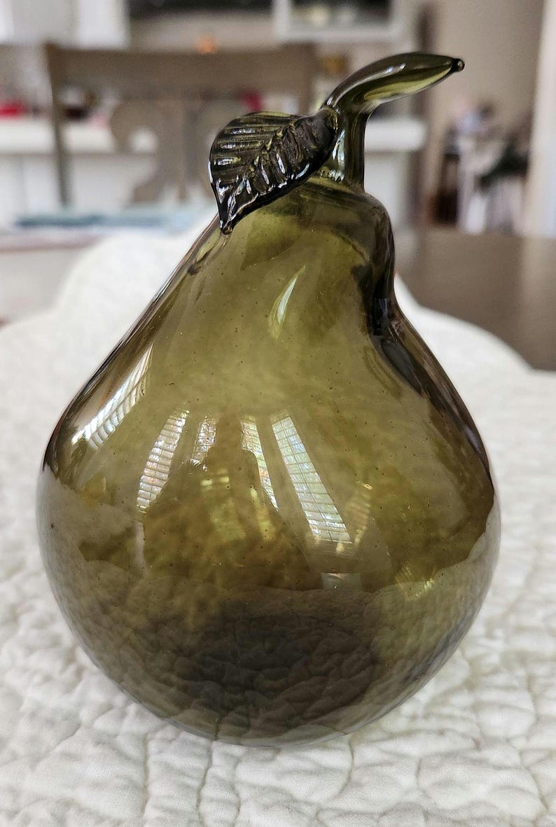 May include: Olive green glass pear-shaped decorative object. The pear has a textured surface and a stem with a leaf detail. The glass is translucent, allowing light to pass through. This decorative piece is suitable for home decor.