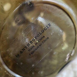 May include: Close-up of a metallic object, possibly brass, with a circular shape. The text "by ANTHROPOLOGIE" is printed in black, along with "Made in China" and "Fabriqu&eacute; en Chine". The object has a reflective surface.