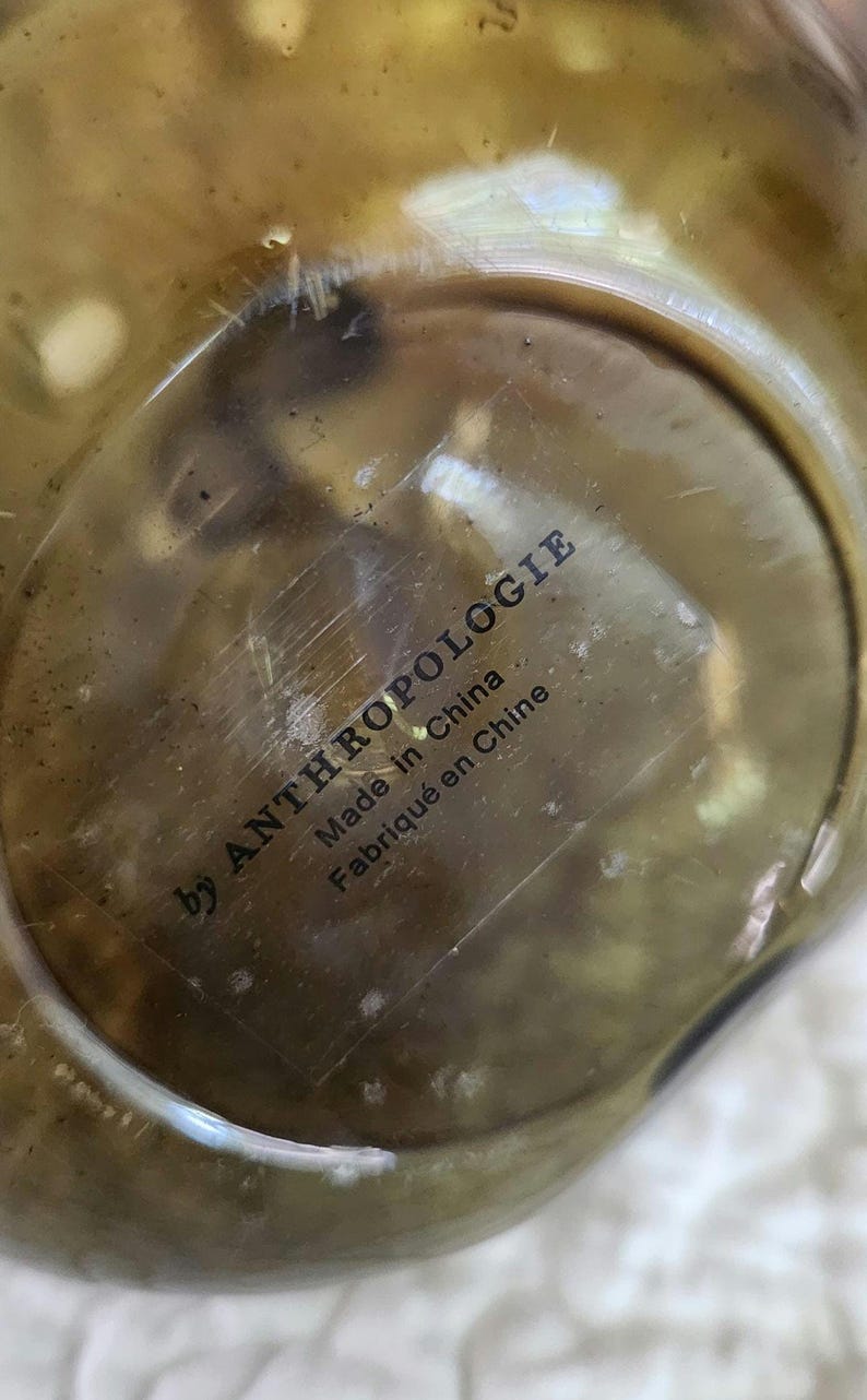 May include: Close-up of a metallic object, possibly brass, with a circular shape. The text "by ANTHROPOLOGIE" is printed in black, along with "Made in China" and "Fabriqu&eacute; en Chine". The object has a reflective surface.
