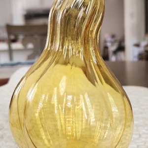 May include: A decorative, amber-colored glass gourd-shaped vase. The vase has a textured surface with vertical ridges and a curved stem. The gourd is sitting on a white, textured surface.