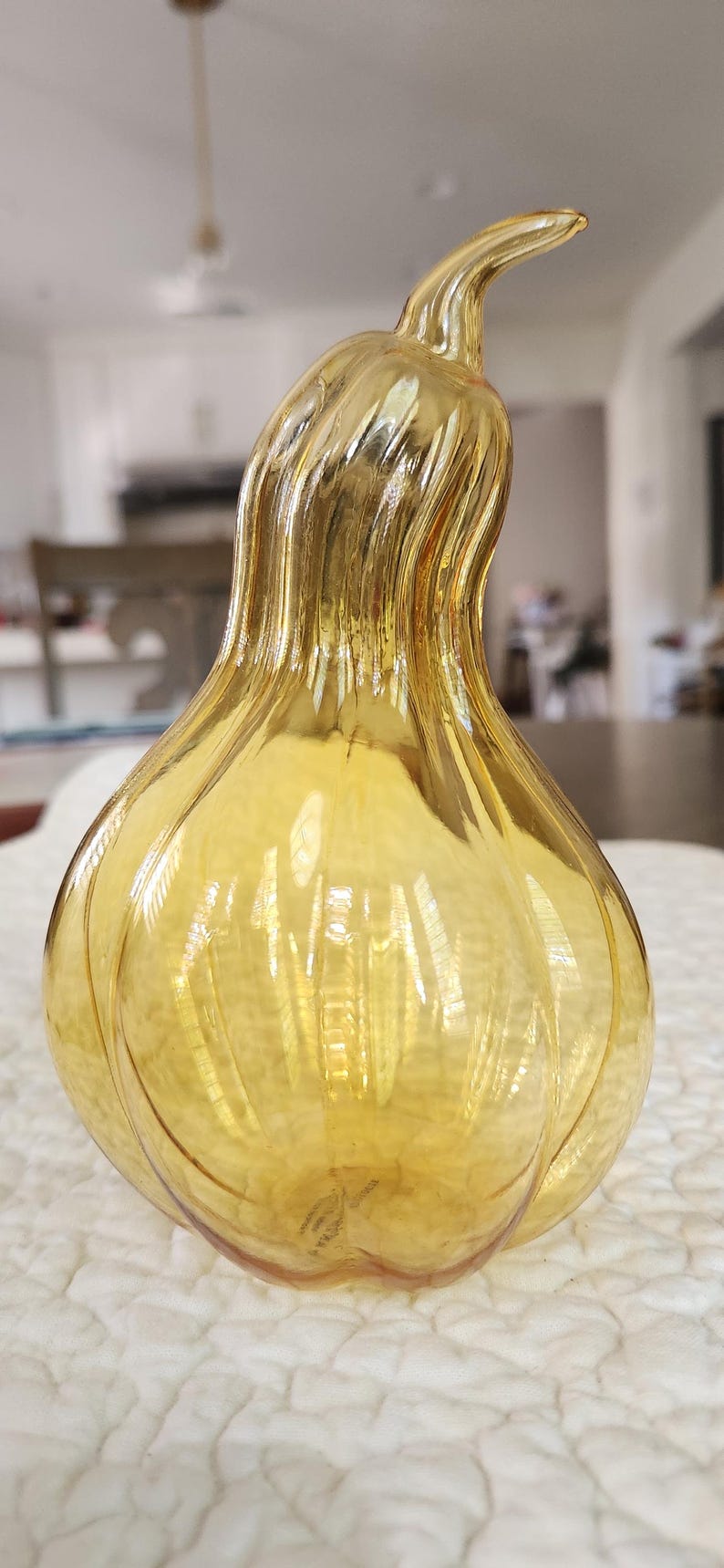 May include: A decorative, amber-colored glass gourd-shaped vase. The vase has a textured surface with vertical ridges and a curved stem. The gourd is sitting on a white, textured surface.