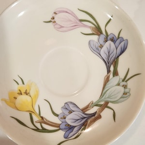 May include: White ceramic saucer with a floral design featuring yellow, purple, and blue crocus flowers. The flowers are arranged around the edge of the saucer with green leaves and brown stems. The saucer has a slightly raised rim.