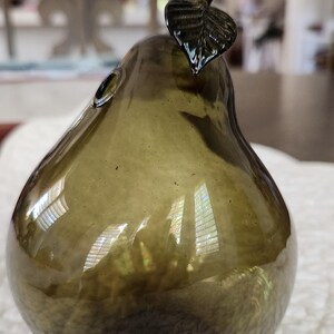 May include: An olive green glass pear ornament. The pear-shaped figurine has a textured surface and a dark green stem and leaf. The decorative object is designed to resemble a pear.