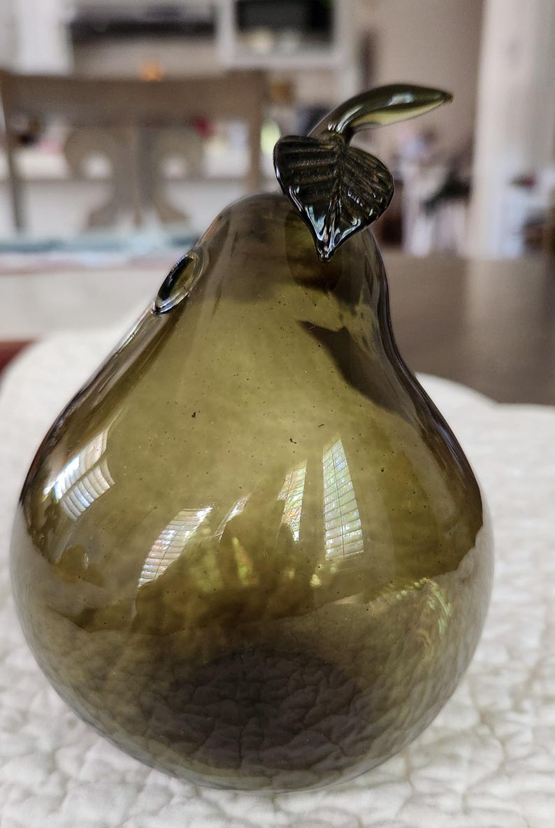 May include: An olive green glass pear ornament. The pear-shaped figurine has a textured surface and a dark green stem and leaf. The decorative object is designed to resemble a pear.