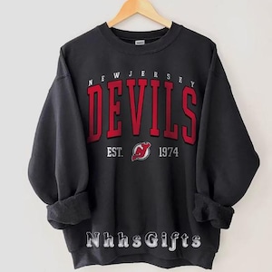 May include: Black sweatshirt with the text "NEW JERSEY DEVILS" in red block letters. Below the text is "EST. 1974" with a red and white logo. The sweatshirt is hanging on a wooden hanger.