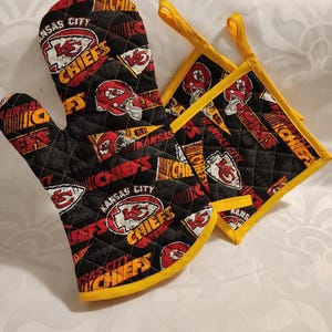 May include: A set of two black and red Kansas City Chiefs oven mitts with yellow trim. The mitts feature a quilted design and the team's logo.