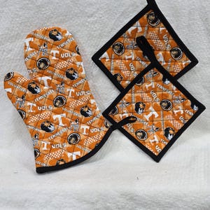 May include: An orange quilted oven mitt and two square pot holders featuring a repeating pattern of the Tennessee Volunteers logo, the letter T, and a dog mascot. Each item is finished with black fabric trim and includes a black hanging loop.