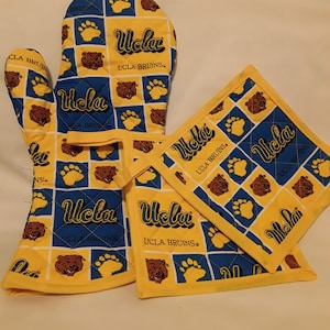 May include: A set of two yellow and blue oven mitts with a UCLA Bruins logo and paw print design. The mitts are quilted and have a yellow border.