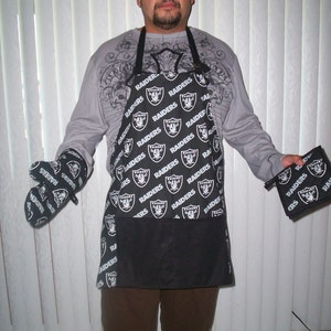 May include: A black and white apron with the word "RAIDERS" repeated across the front. The apron has a large pocket on the front. The person wearing the apron is also wearing black and white oven mitts.