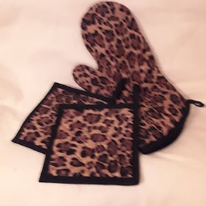 May include: A set of leopard print kitchen accessories, including a potholder and two hot pads. The potholder has a black trim and a loop for hanging. The hot pads are square and have black trim.
