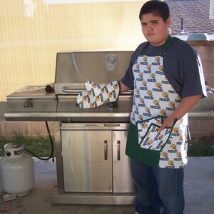 Op de afbeelding: Een persoon draagt een groen-witte schort met het woord "Packers" dat erop herhaald wordt. De schort heeft een groene zak aan de voorkant. De persoon staat voor een roestvrijstalen grill met een propaantank aan de linkerkant.