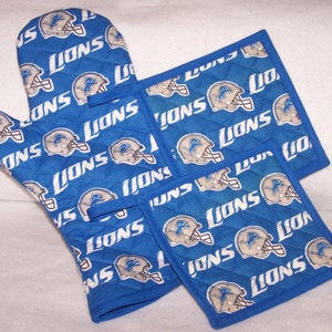 May include: A set of blue kitchen pot holders with a white quilted pattern featuring the Detroit Lions logo and the word "Lions" repeated throughout.