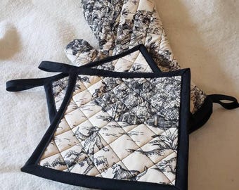 May include: A set of two black and white quilted oven mitts with a black trim. The mitts feature a black and white toile pattern with a repeating design of a house, trees, and people.