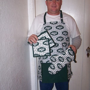 May include: A man wearing a white shirt with a green and white apron with the word "Jets" repeated on it. He is holding a set of green and white oven mitts and pot holders with the word "Jets" repeated on them.