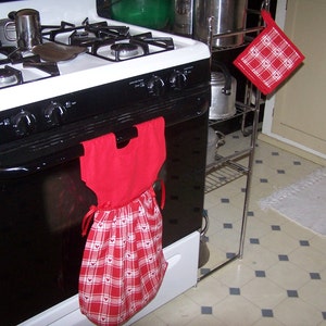 May include: A red and white plaid apron with heart accents hangs from the handle of a black oven. A matching red pot holder with white heart details hangs on a nearby metal rack. A white and black stove top is visible.