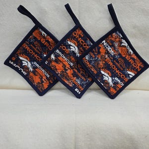 Op de afbeelding: Drie vierkante, doorgestikte pannenlappen met een patroon van het Denver Broncos teamlogo en tekst in marineblauw, oranje en wit. Elke pannenlap heeft een donkerblauwe stoffen rand en een bijpassend stoffen lusje aan een hoek om op te hangen.