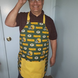 May include: A green and yellow apron with the Green Bay Packers logo repeated all over. The apron has a large yellow pocket on the front.