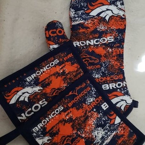 Puede incluir: Un juego de dos manoplas para horno azules y naranjas con un diseño acolchado. Las manoplas presentan un patrón repetido del logotipo de los Denver Broncos y las palabras "Broncos" y "Denver".
