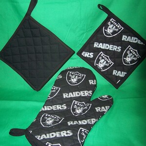 May include: Set of three black quilted oven mitts with a white Raiders logo and text pattern. The mitts include two square pot holders and one oven mitt.